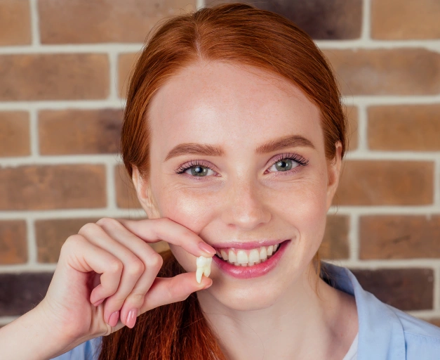 Young woman with red hair smiling, pointing to teeth against brick wall background.