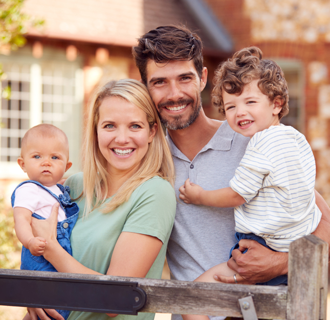 Happy family of four smiling together outdoors in front of brick home.